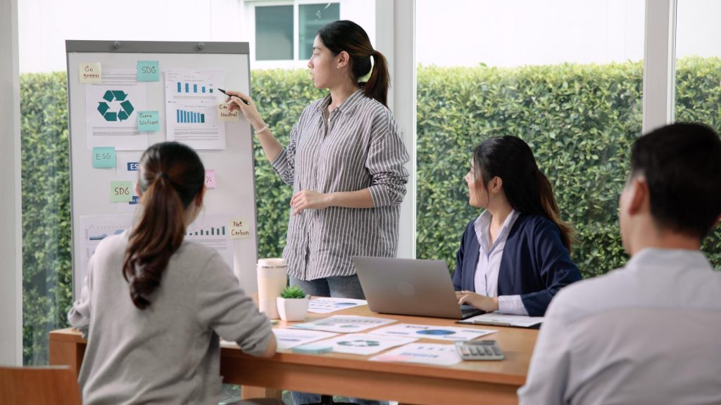 Schreibtisch mit Dokumenten und einem Laptop, zwei Frauen schauen zu Whiteboard, neben dem eine Frau steht, MKC Martin Kaufmann Consultation